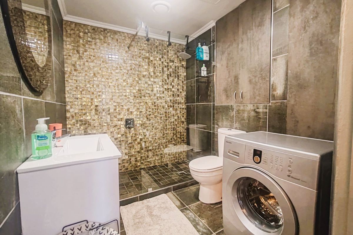 A modern bathroom featuring a glass-enclosed walk-in shower, mosaic tile accent wall, vanity basin, toilet, and integrated laundry space with a washing machine.