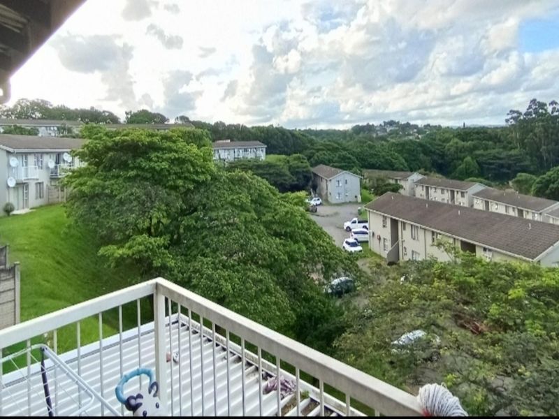 A view from an elevated balcony overlooking a residential apartment complex with surrounding greenery and parking areas.