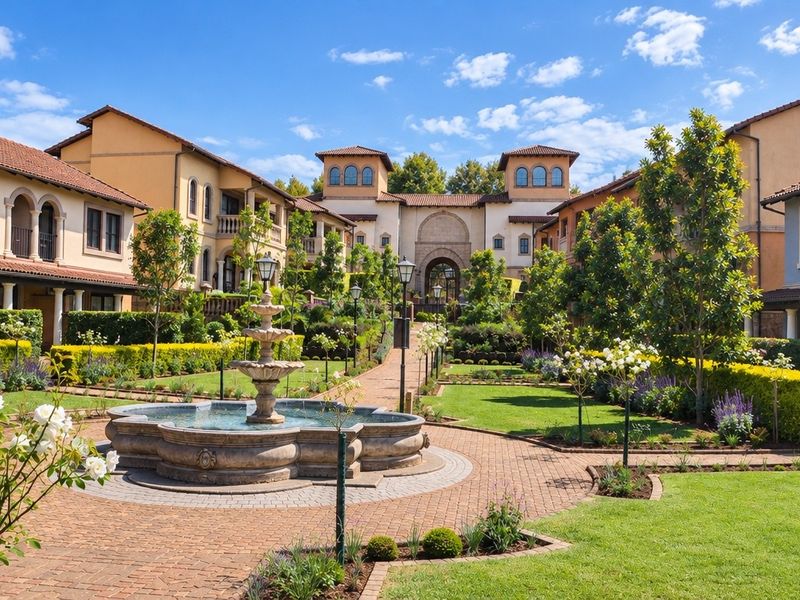 A beautifully landscaped central courtyard featuring a stone fountain, brick pathways, and well-maintained gardens surrounded by Mediterranean-style apartment buildings.