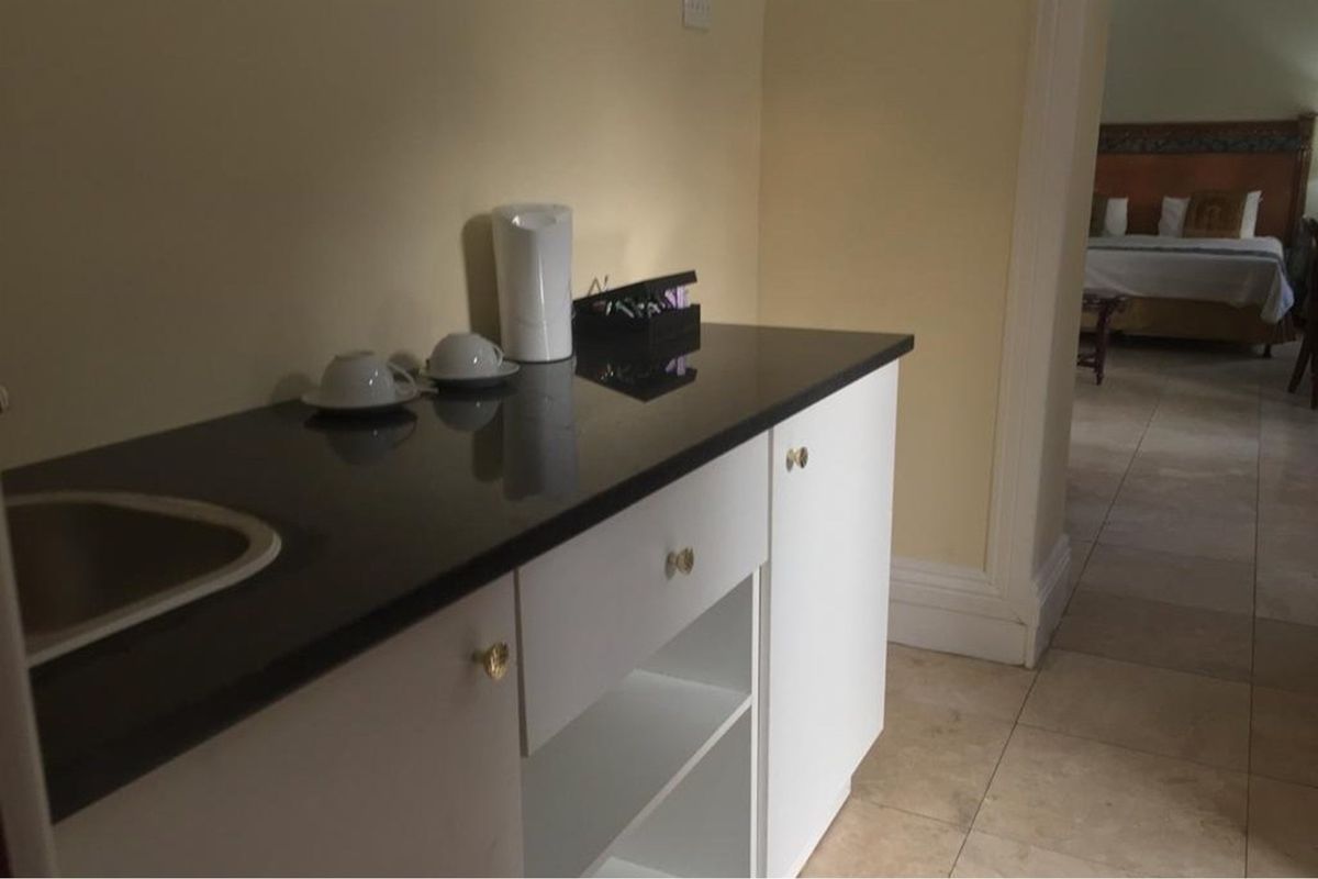 A compact kitchenette area featuring white cabinetry, a dark countertop, and an integrated sink, with a view into the adjacent bedroom.