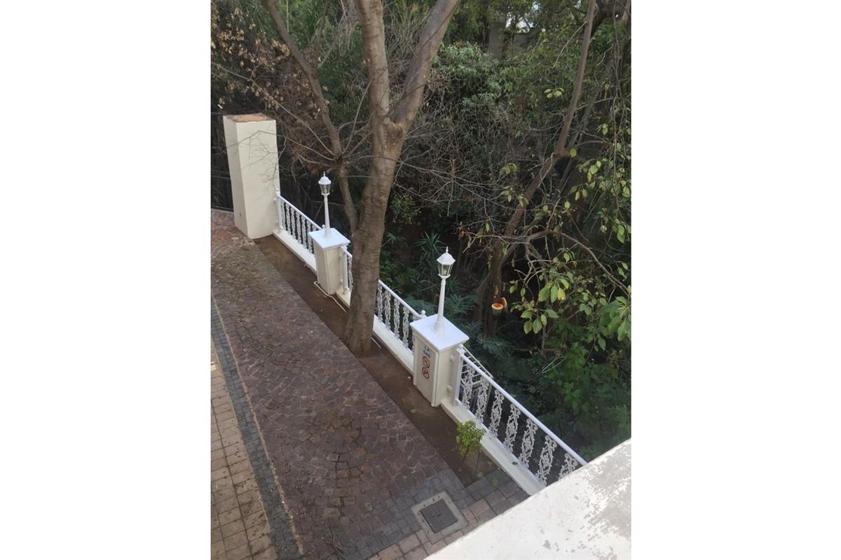 An elevated view of a paved walkway featuring decorative white railings, integrated lamp posts, and mature trees overlooking a garden area.