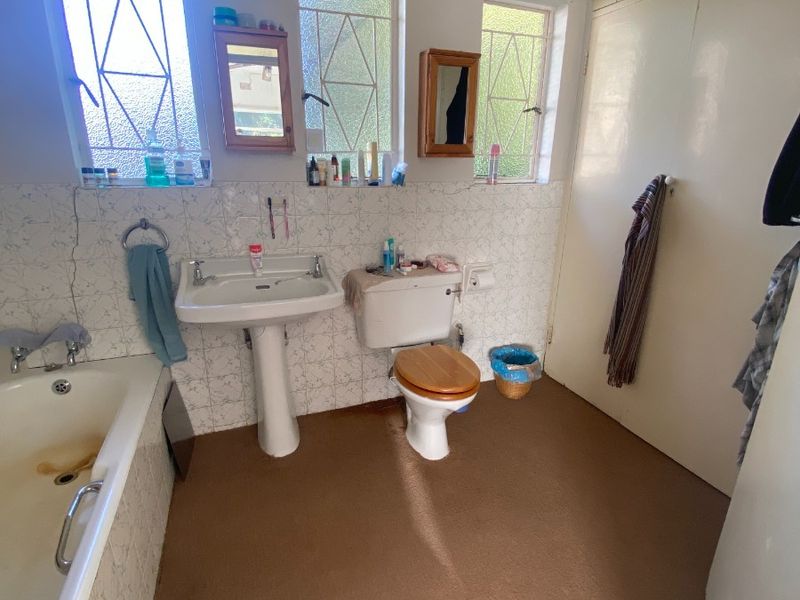 A dated bathroom featuring a pedestal basin, toilet, and bathtub, with built-in storage cupboards and natural light from three windows.