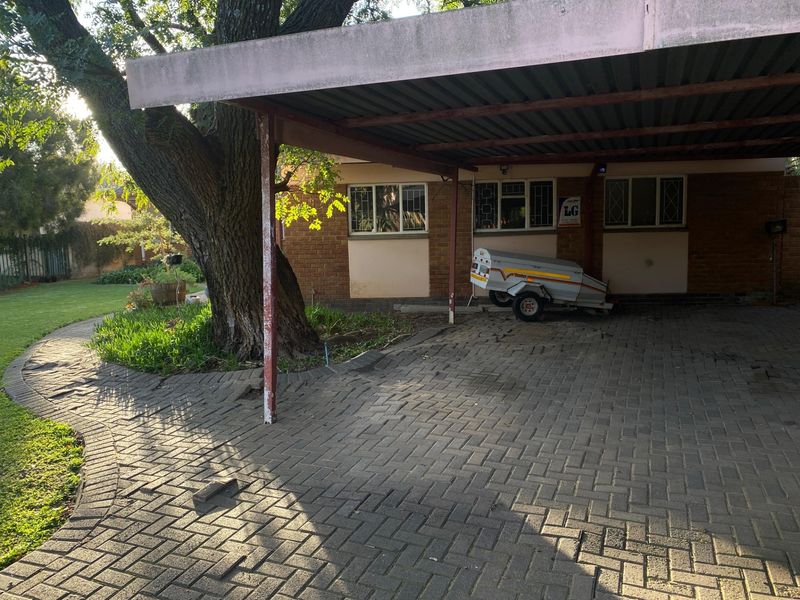 Front exterior view featuring a paved driveway, covered carport, and established garden with mature trees.