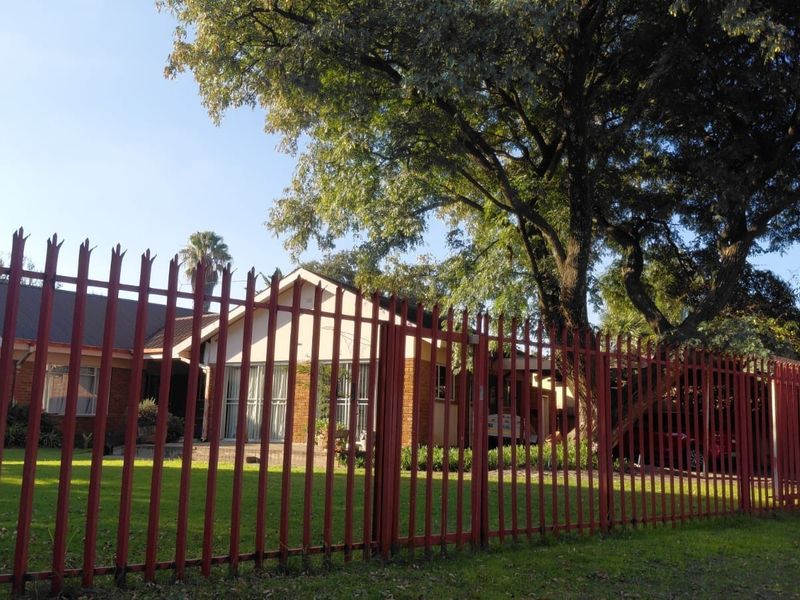 Front view of a brick house featuring a large mature tree, well-maintained lawn, and secure steel palisade fencing.