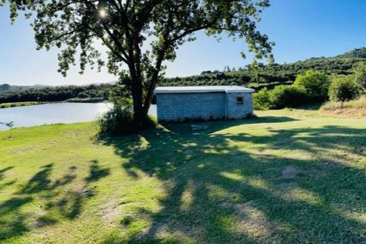 A small outbuilding situated on a grassy farm plot overlooking a dam, surrounded by mature trees and natural landscape.