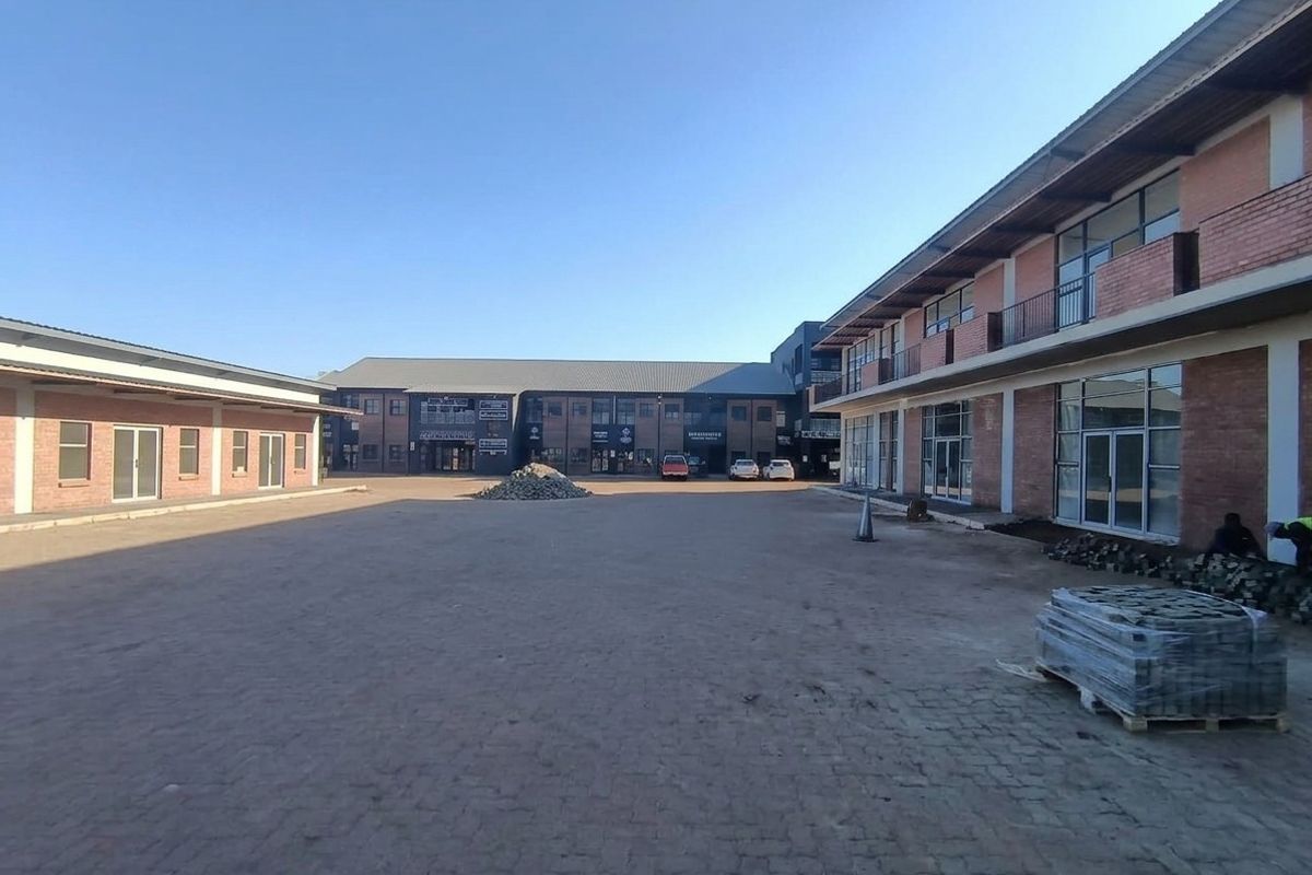 A commercial courtyard featuring brick buildings with glass storefronts, balconies, and paved ground, currently undergoing final site finishing.