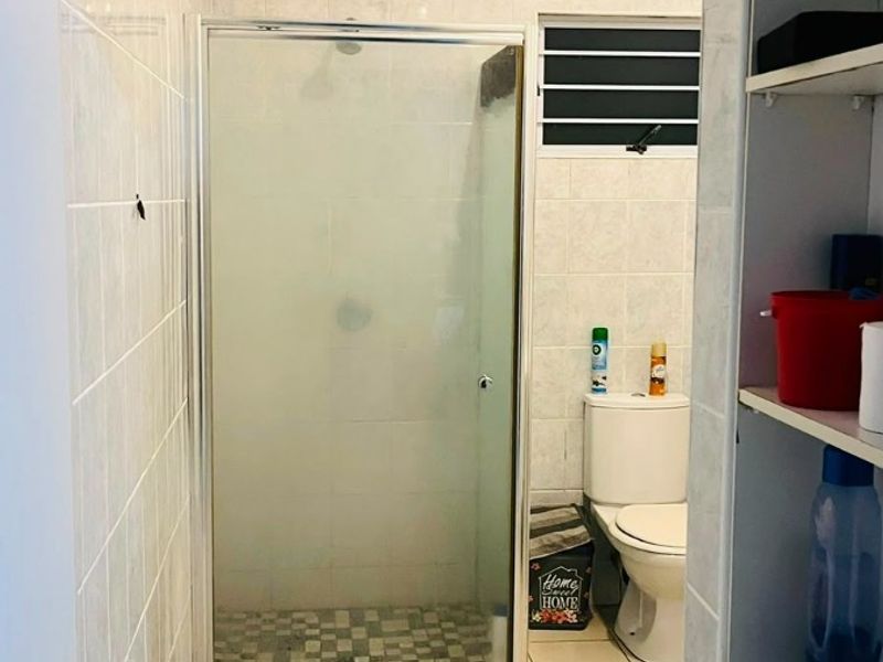 A functional bathroom featuring a glass-enclosed shower, tiled walls and flooring, and a standard toilet, viewed from an adjacent storage area.