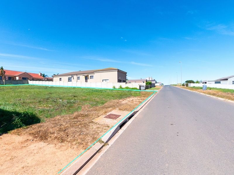 A street-level view of a vacant residential plot featuring established asphalt road access, concrete curbing, and existing boundary walling in a developed neighborhood.