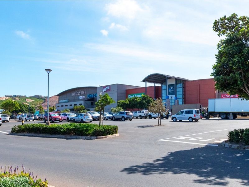 View of the Weskus Mall commercial property featuring a large paved parking area, landscaped garden beds, and modern building facade.