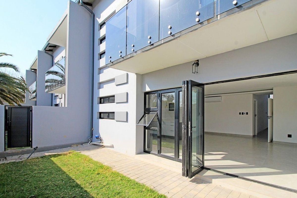 Modern rear exterior view featuring a paved patio, glass balcony balustrade, and large bi-fold doors leading into the open-plan living space.
