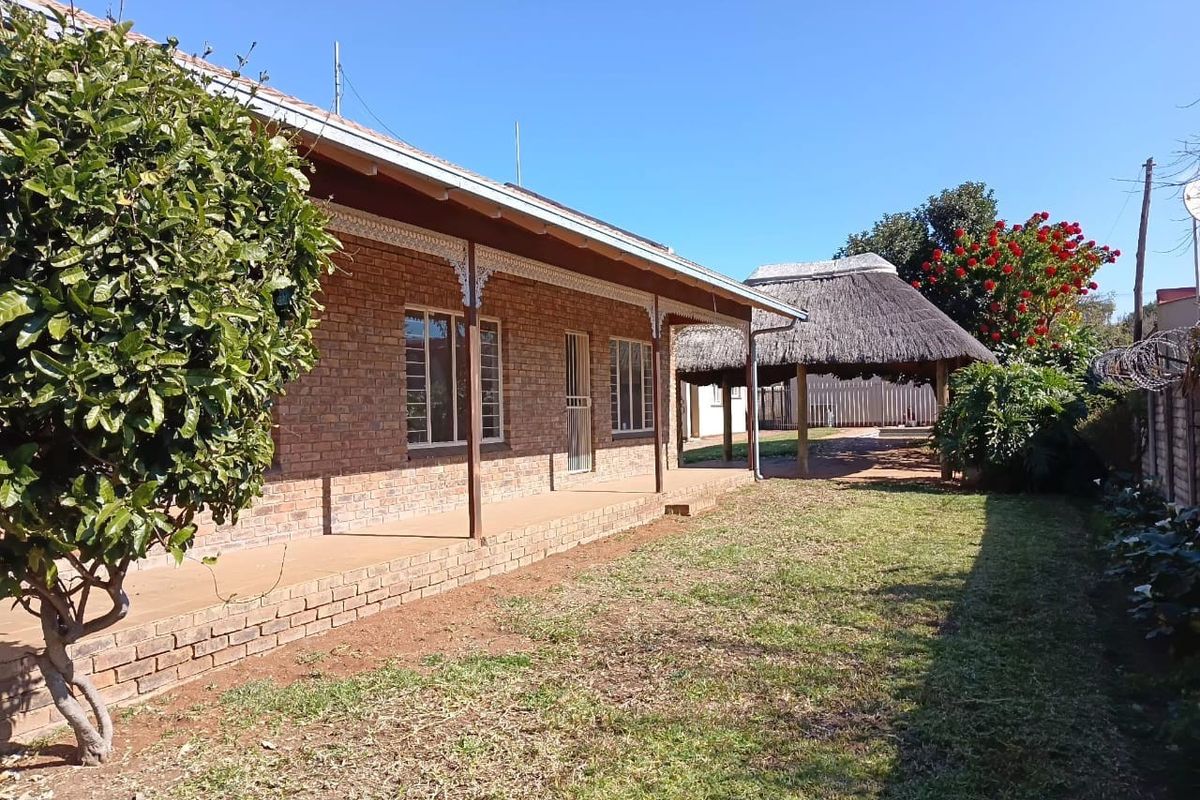 Rear view of the house featuring a brick facade, a covered veranda, and a detached thatched lapa set within a garden area.