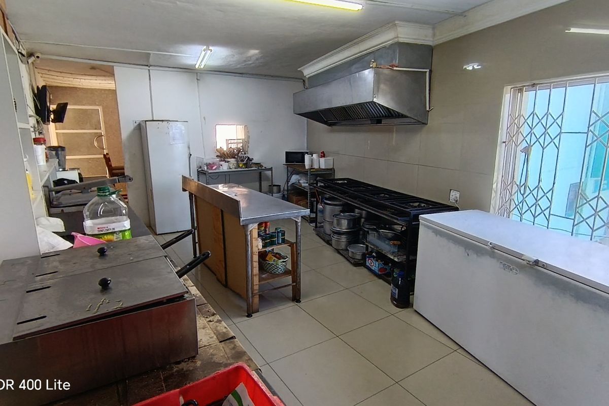 Commercial kitchen featuring stainless steel prep tables, industrial extractor hood, tiled flooring, and security bars on the window.
