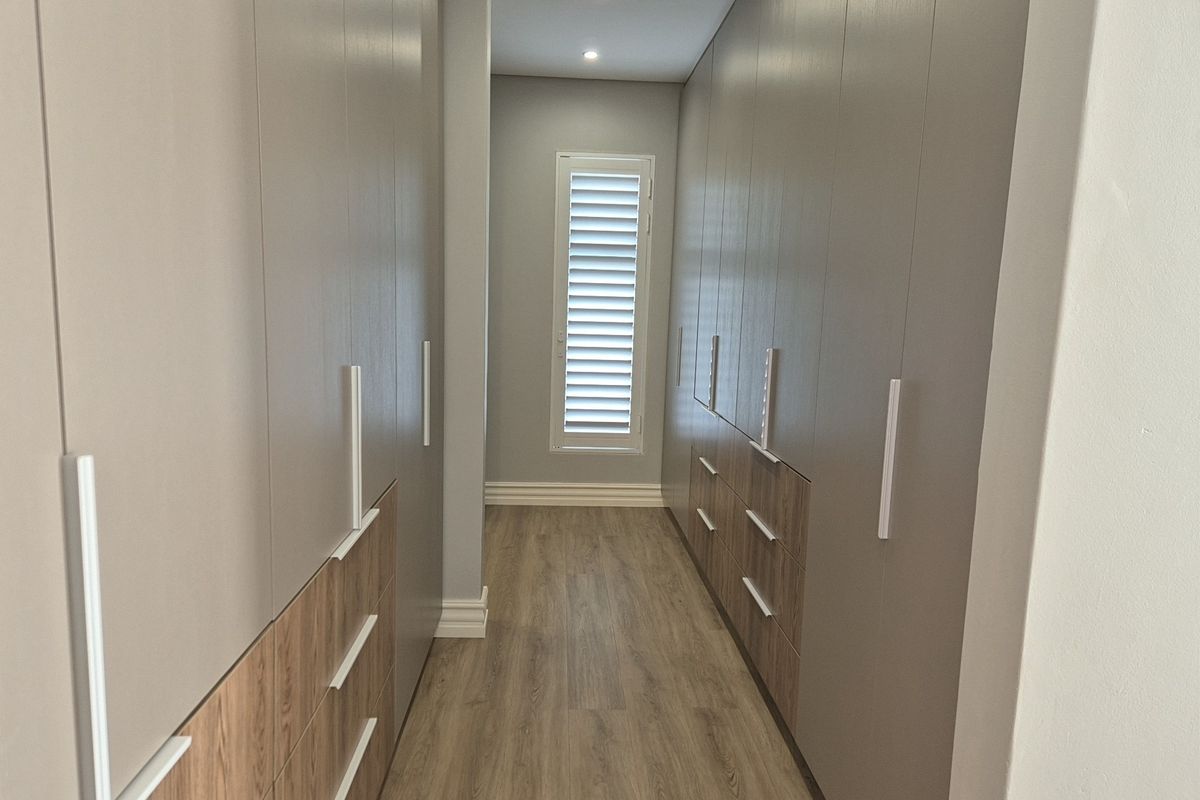 A modern walk-in dressing area featuring extensive floor-to-ceiling built-in cabinetry with a mix of wood-grain and neutral finishes, complemented by plantation shutters.