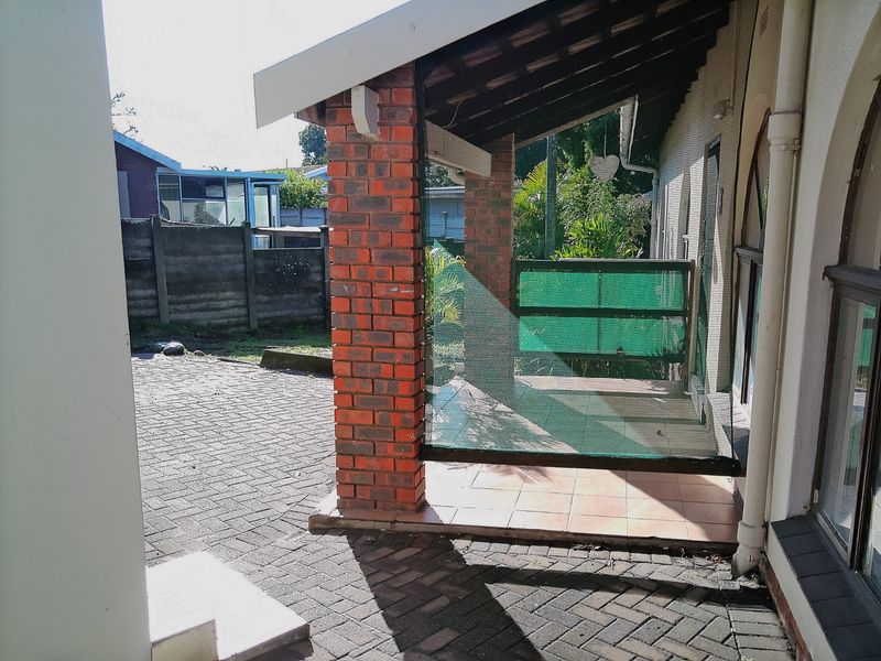 A covered outdoor patio area featuring brick pillars and tiled flooring, adjacent to a paved driveway and rear garden space.