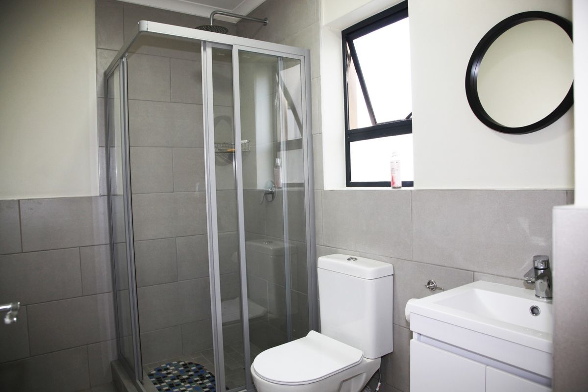 A modern bathroom featuring a glass-enclosed shower, a white vanity with a basin, and a toilet, finished with neutral grey wall tiles.