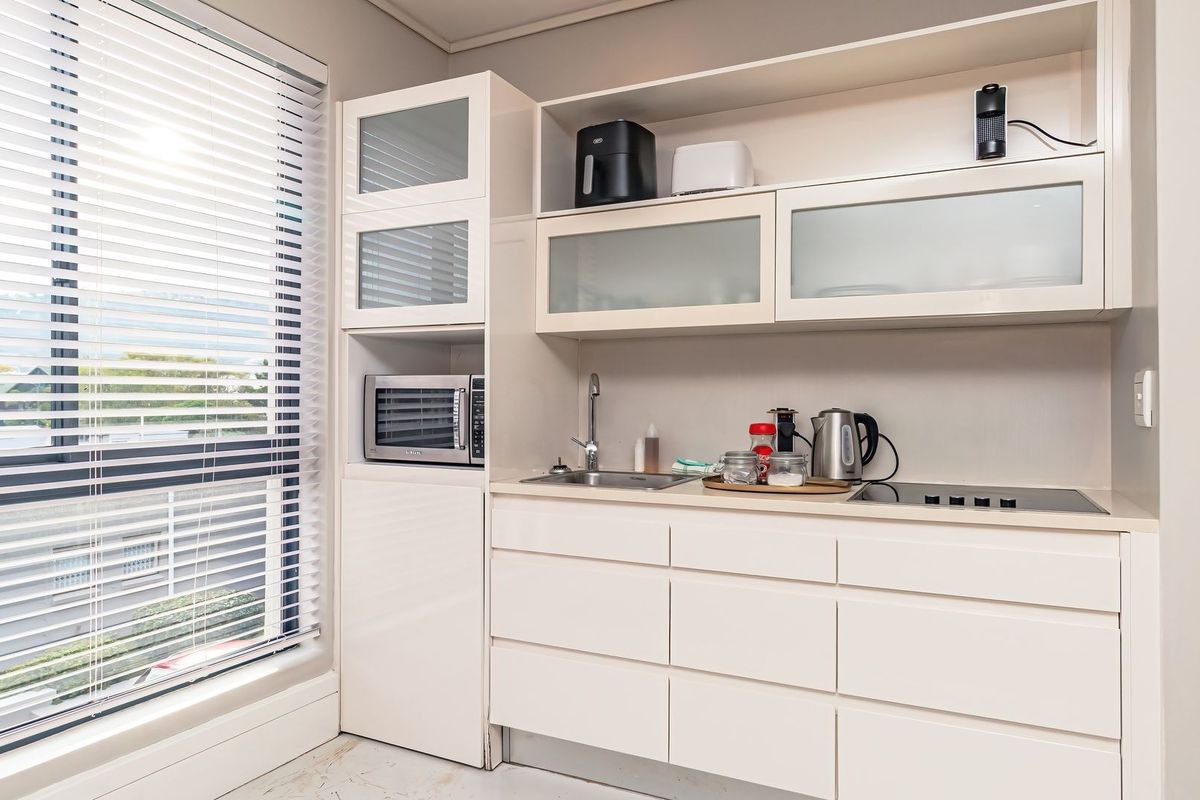 Compact kitchenette featuring white cabinetry, integrated sink, built-in microwave, and a sleek cooktop, positioned next to a large window with fitted blinds.