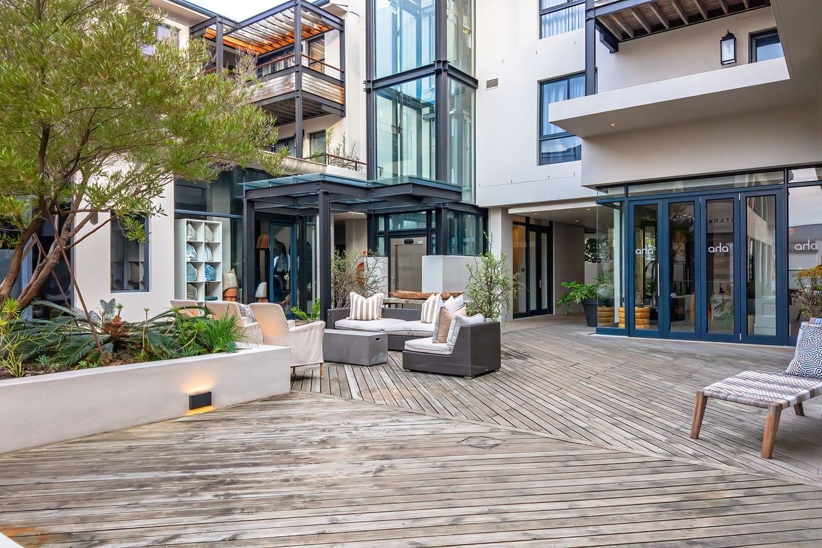A modern apartment complex courtyard featuring timber decking, landscaped garden beds, and glass-enclosed architectural elements.