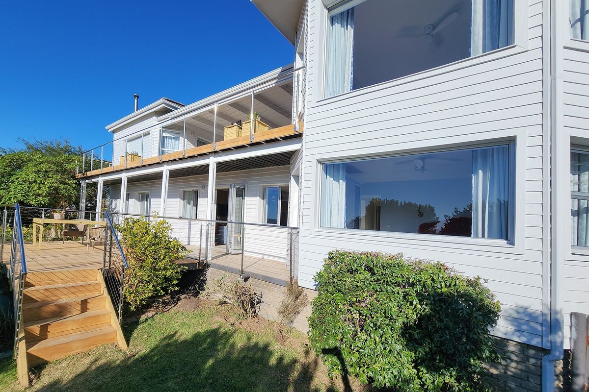 A multi-level exterior rear view of a white weatherboard house featuring a spacious wooden deck, stainless steel railings, and large glass windows.