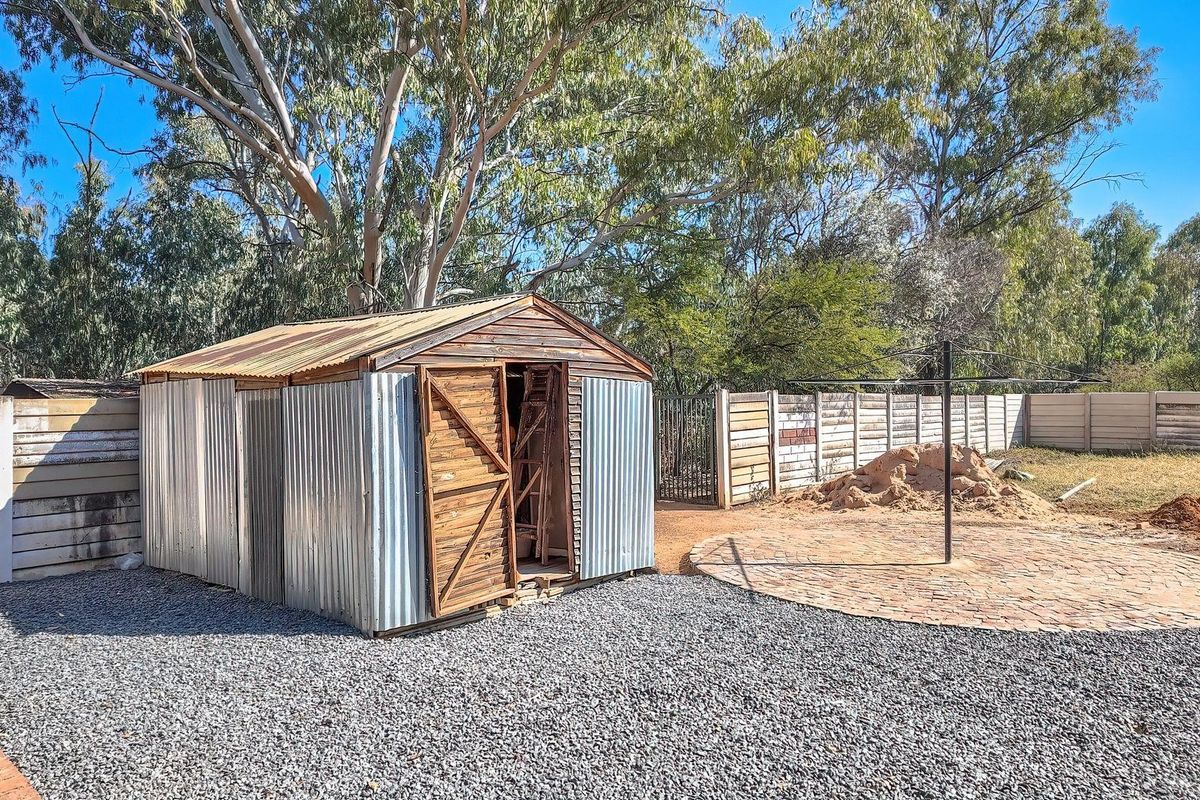 A rustic corrugated metal storage shed situated in a gravel-surfaced backyard with a paved circular area and perimeter boundary walling.