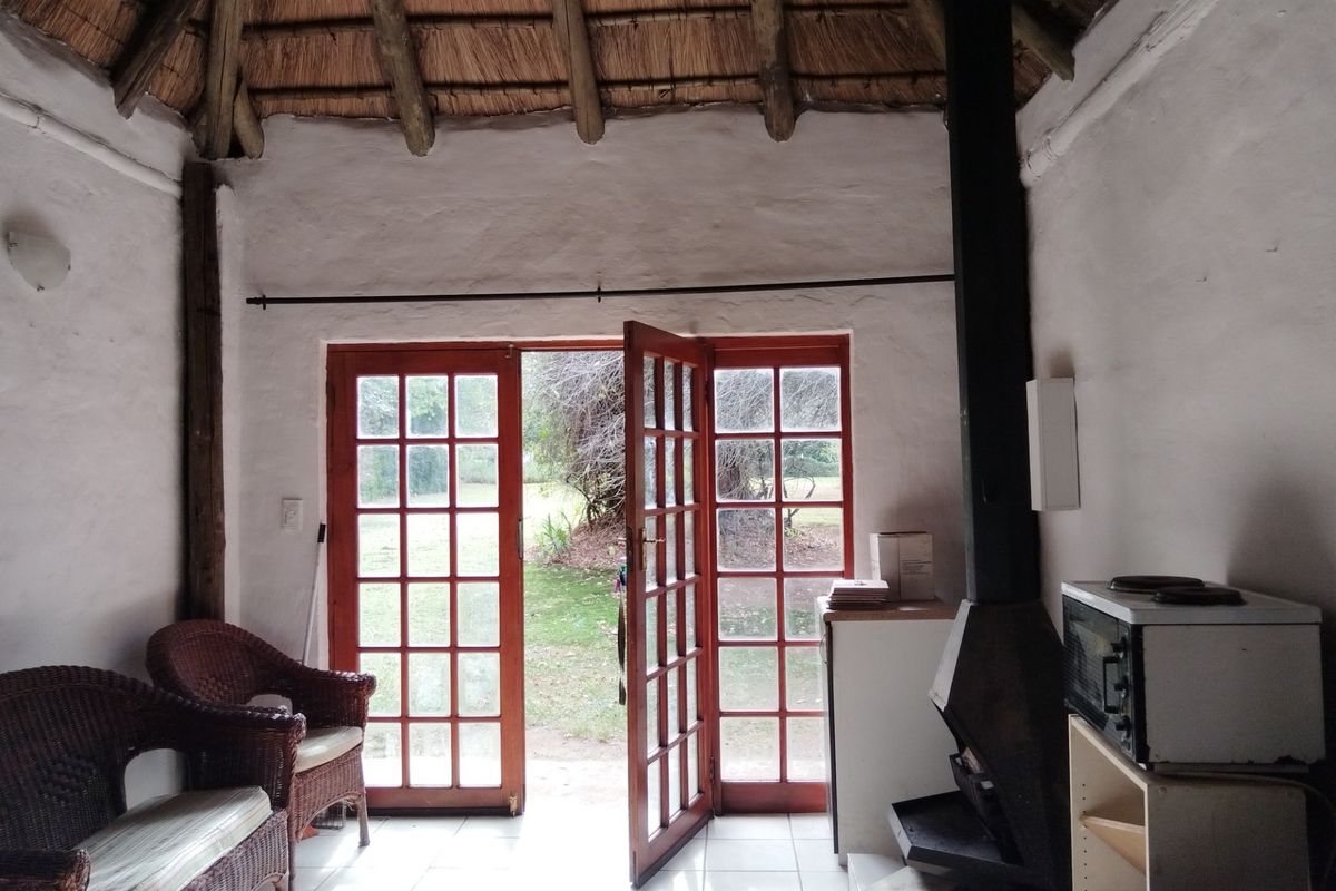 A rustic living area featuring a traditional thatched roof, tiled flooring, and a built-in fireplace, with direct access to the garden through French doors.