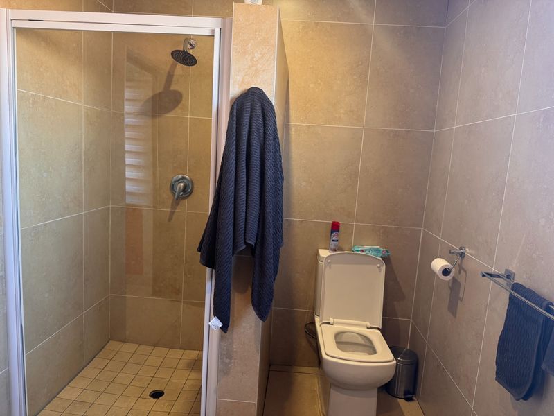 A functional bathroom featuring a walk-in shower with a rainfall shower head, a toilet, and neutral floor-to-ceiling wall tiling.