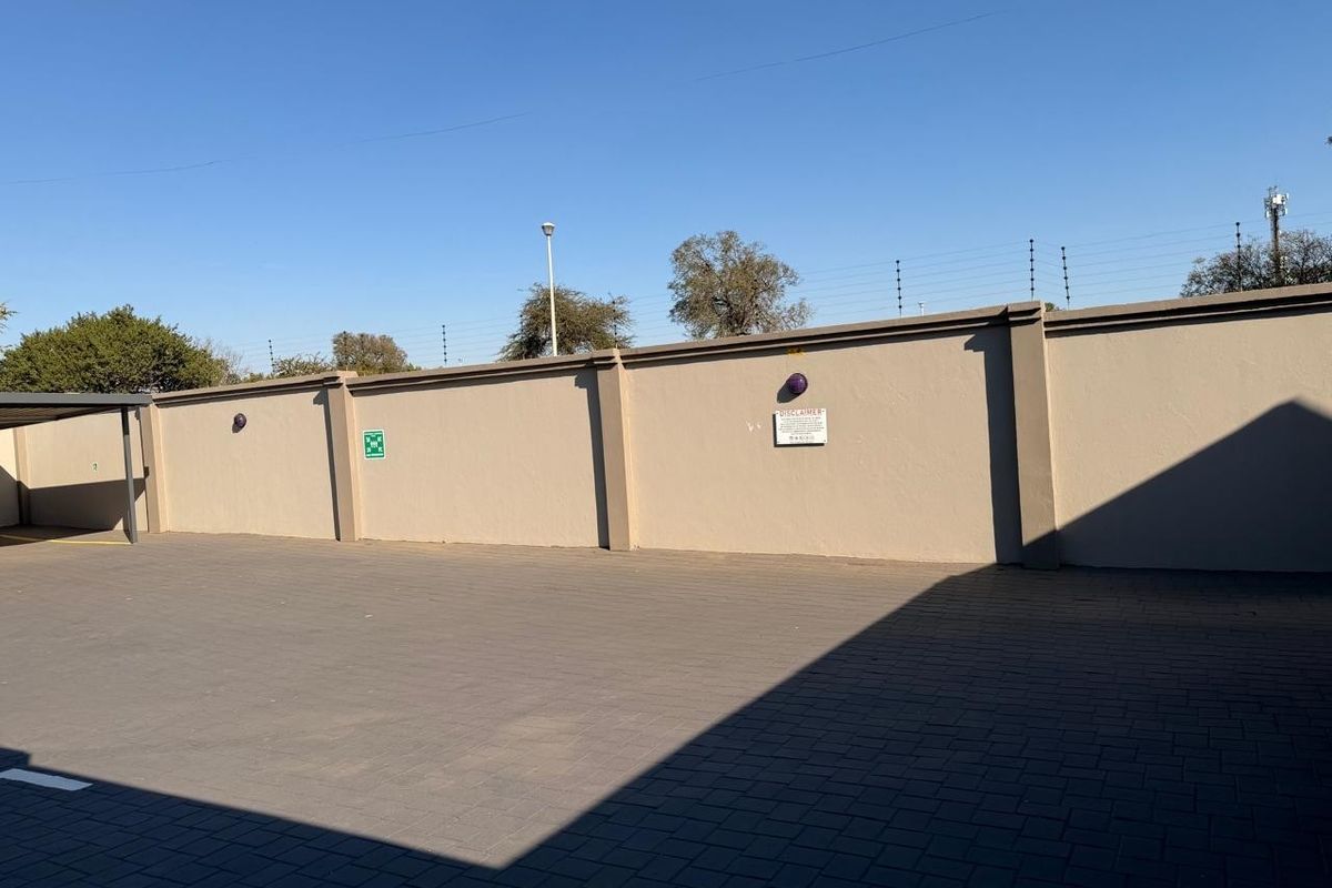 A paved outdoor parking area featuring a high perimeter boundary wall with integrated security fencing and exterior lighting fixtures.