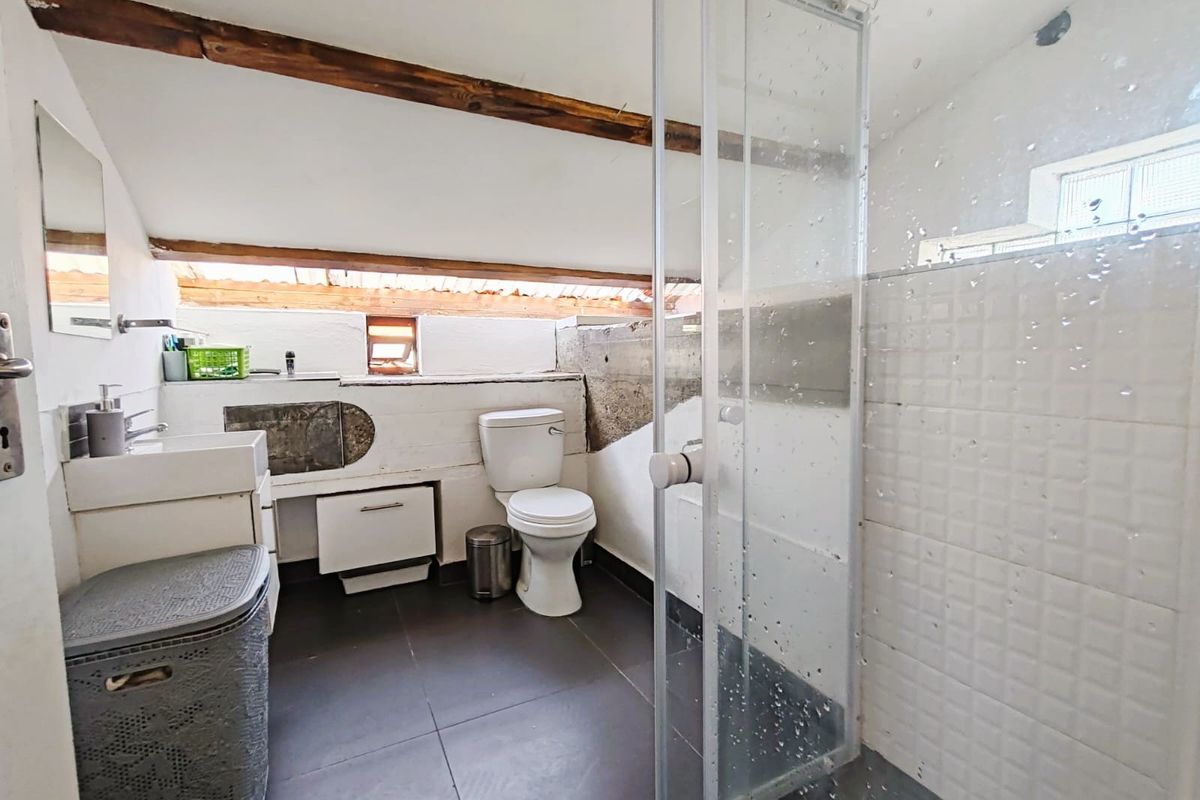 A compact bathroom featuring a glass shower enclosure, toilet, and vanity, characterized by exposed wooden ceiling beams and unfinished wall textures.