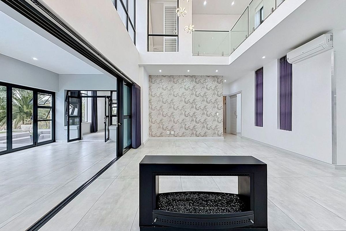 Spacious double-volume living area featuring large glass sliding doors, a modern fireplace, tiled flooring, and a mezzanine level with glass balustrades.