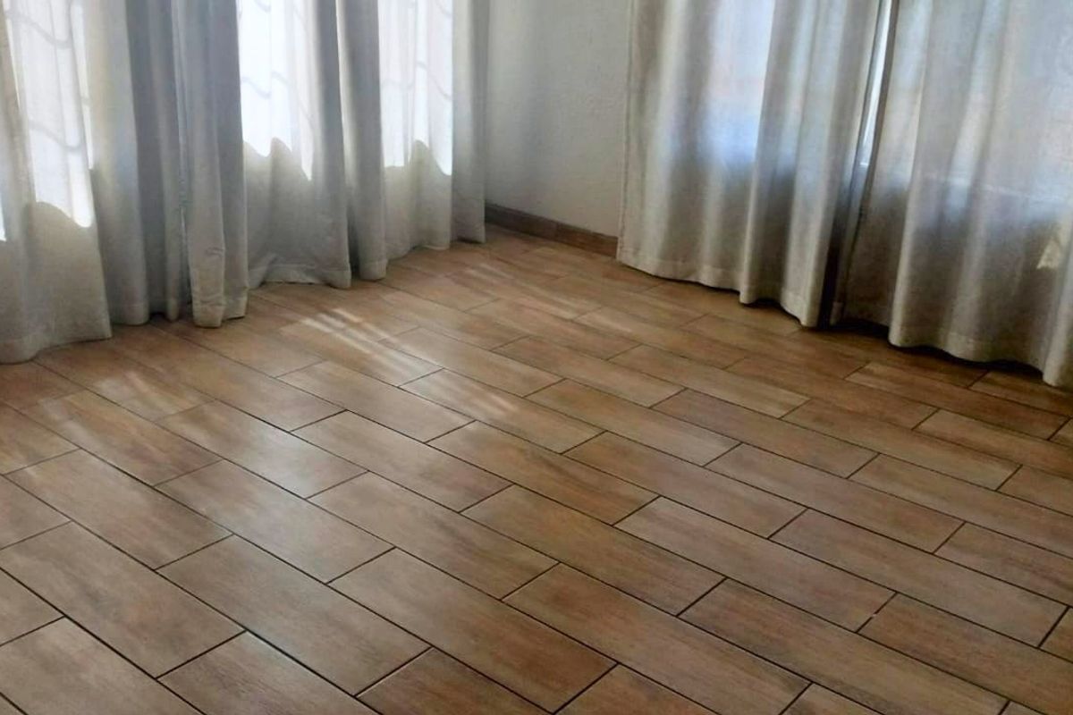 A bright, empty living space featuring modern wood-look ceramic floor tiles and large windows dressed with neutral curtains.