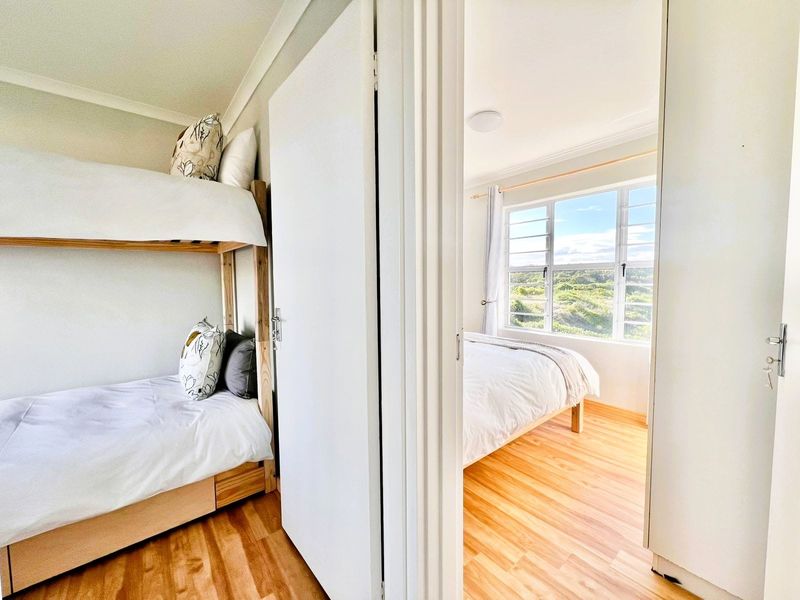 A view through a doorway into a bright bedroom featuring wood-look flooring and large windows with natural light.