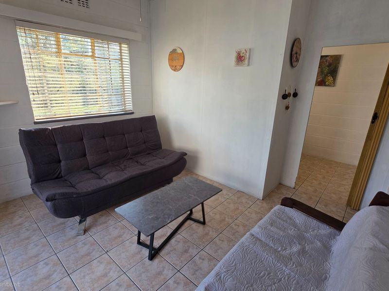 A simple living room featuring tiled flooring, a large window with blinds, and access to an adjacent room.