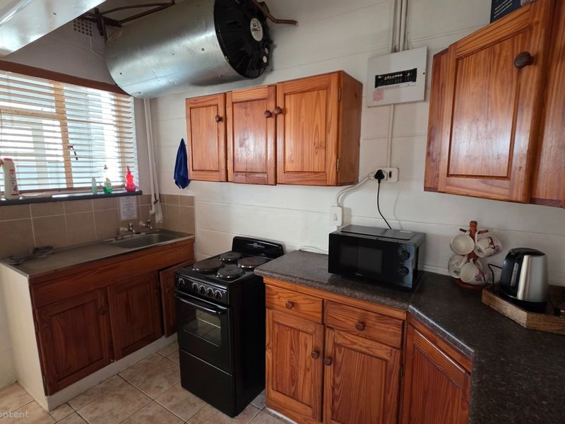 Compact kitchen featuring wooden cabinetry, a freestanding stove, and a stainless steel sink, with exposed ventilation ducting and functional tiled flooring.