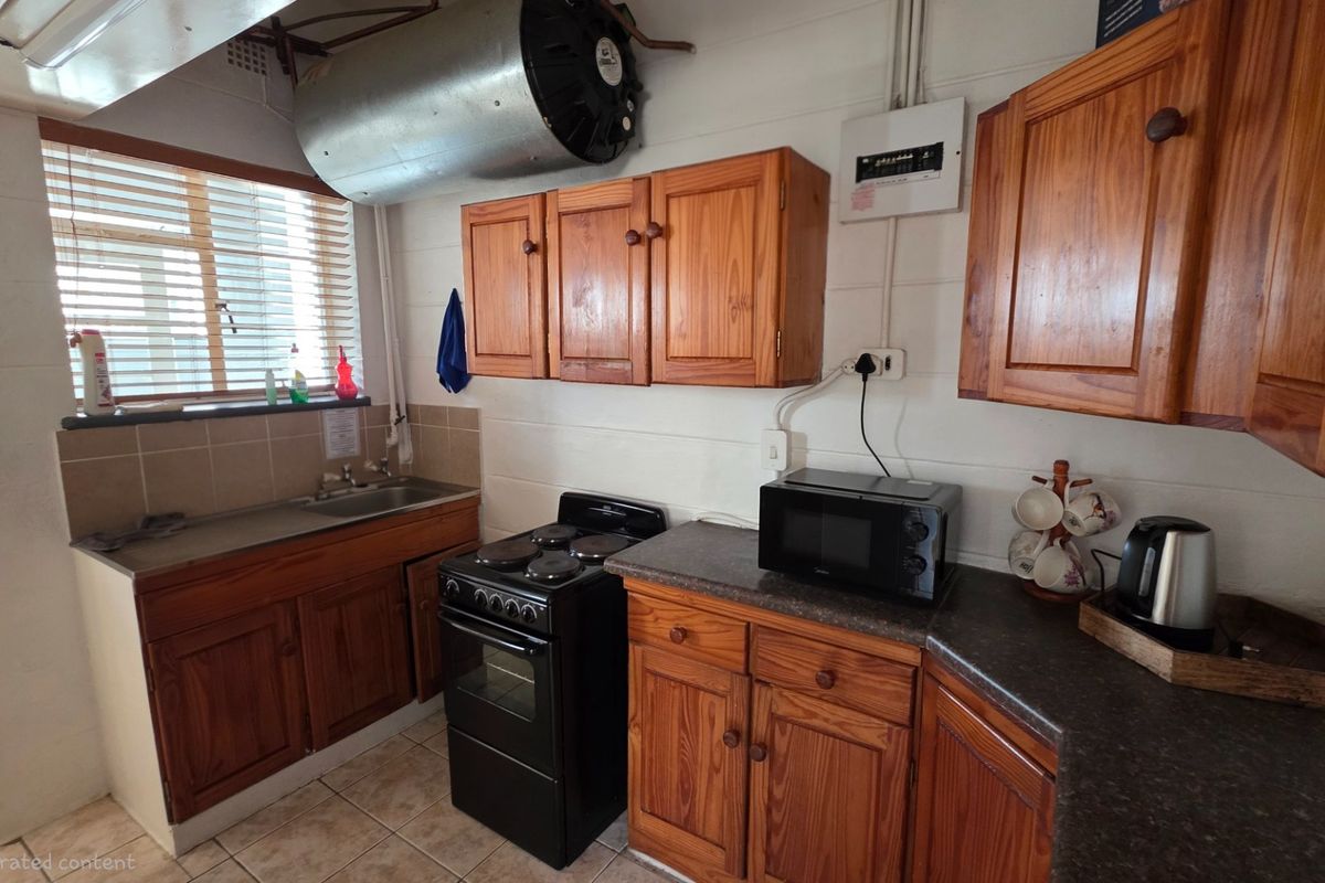 Compact kitchen featuring wooden cabinetry, a freestanding stove, and a stainless steel sink, with exposed ventilation ducting and functional tiled flooring.