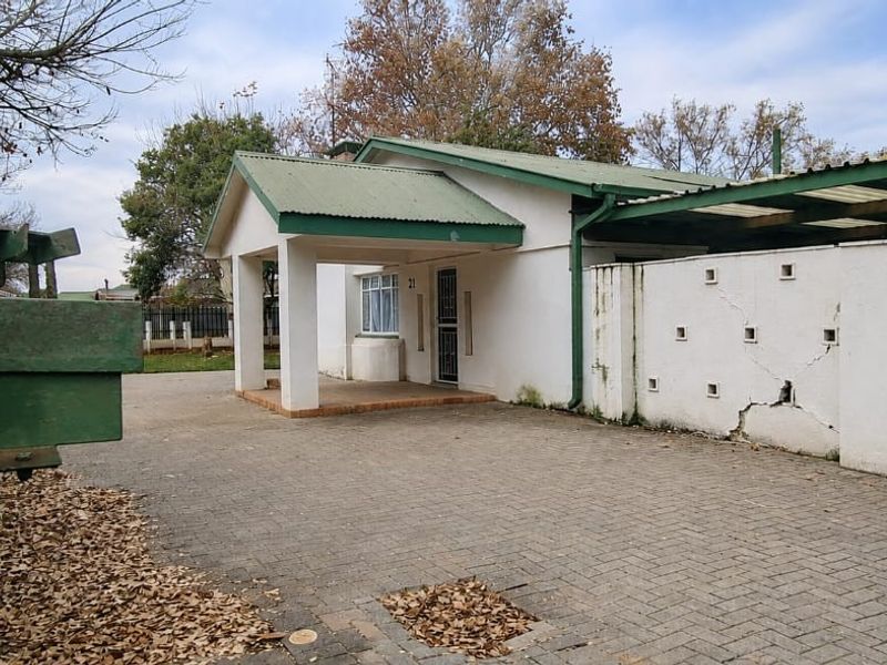 Front exterior view of a single-story house featuring a paved driveway, covered entrance porch, and a carport with a boundary wall showing structural cracks.
