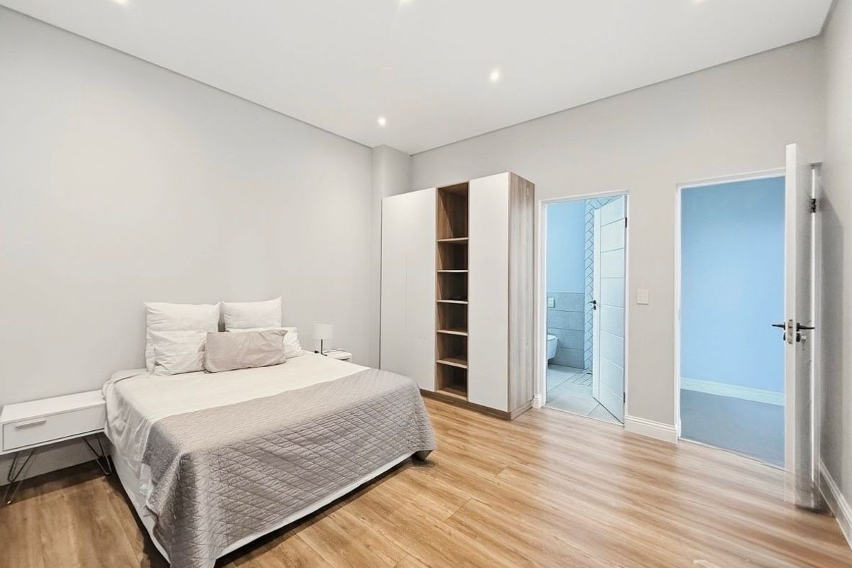 A bright, modern bedroom featuring light wood-look flooring, recessed ceiling lighting, and a built-in wardrobe with open shelving, leading to an en-suite bathroom.