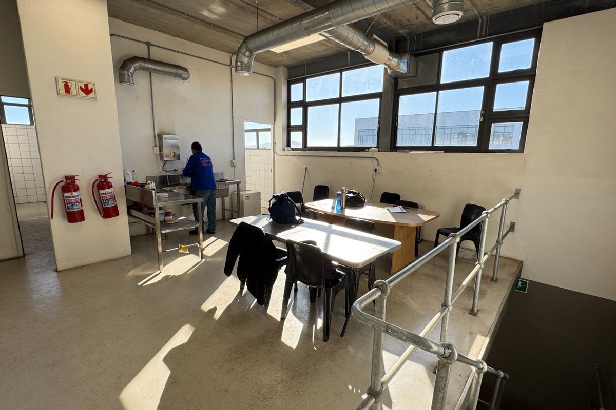 Industrial break room featuring a kitchenette area, exposed concrete ceiling, large windows, and metal safety railing overlooking a stairwell.