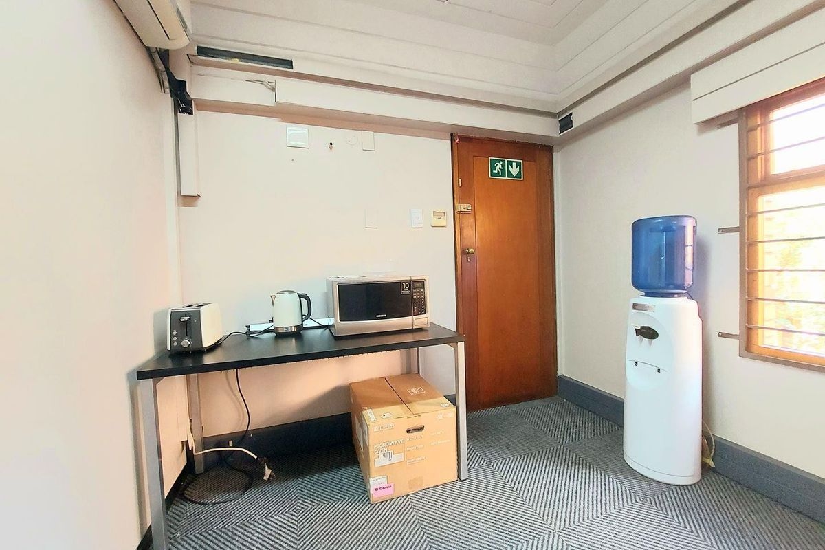 A functional office kitchenette area featuring a wall-mounted air conditioning unit, carpeted flooring, and a wooden door with emergency exit signage.