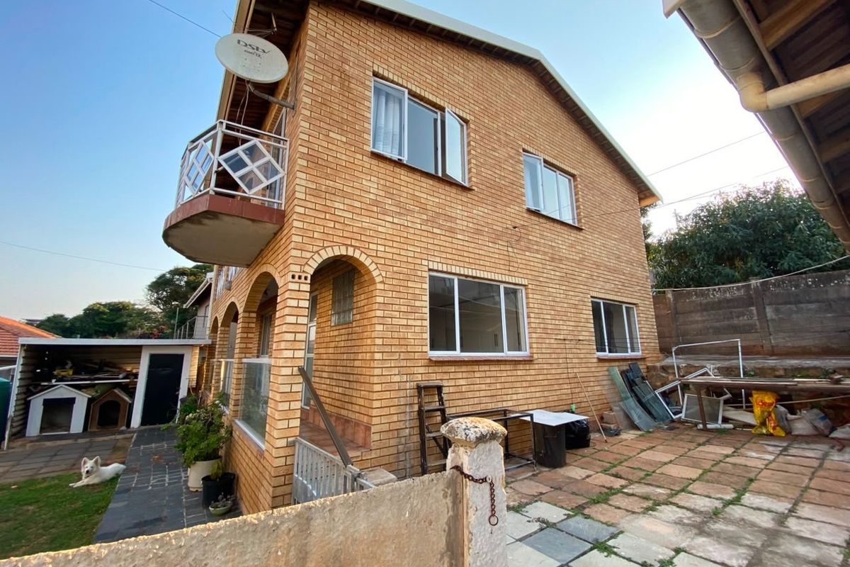 Exterior view of a double-story brick apartment building featuring a small balcony, tiled patio area, and surrounding garden space.