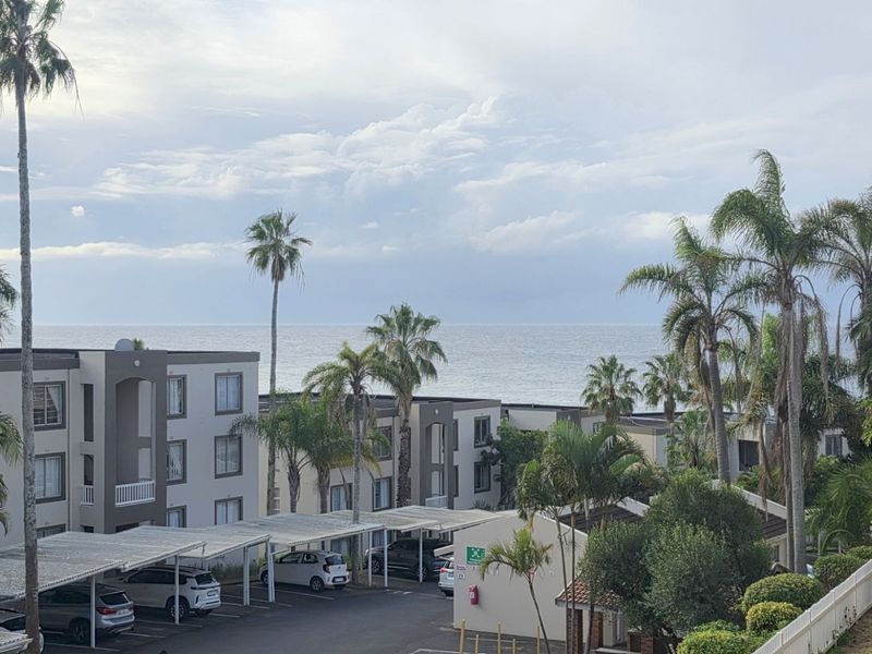 Elevated view overlooking the apartment complex grounds, featuring covered parking bays, landscaped palm trees, and a scenic ocean horizon in the background.