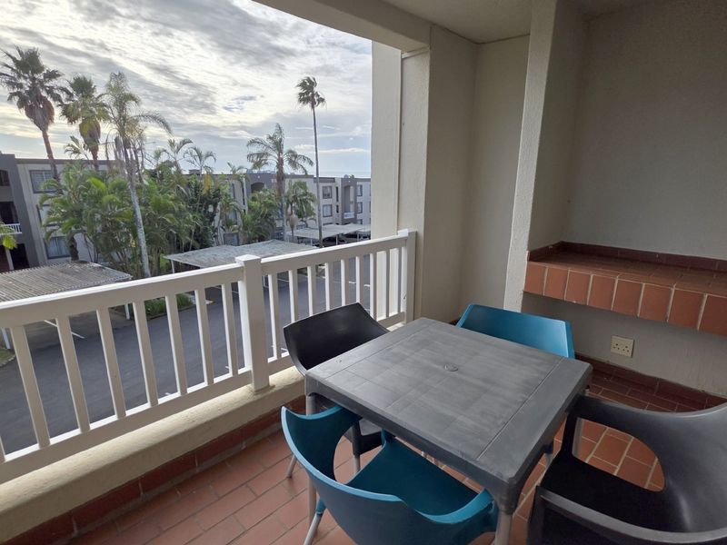 Covered balcony featuring a built-in brick braai, tiled flooring, and white balustrade railing, offering views over the apartment complex grounds.
