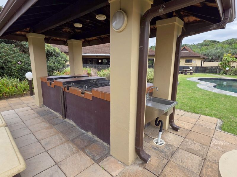 Covered outdoor entertainment area featuring a built-in braai station, stainless steel prep sink, and tiled flooring, overlooking the communal garden and swimming pool.