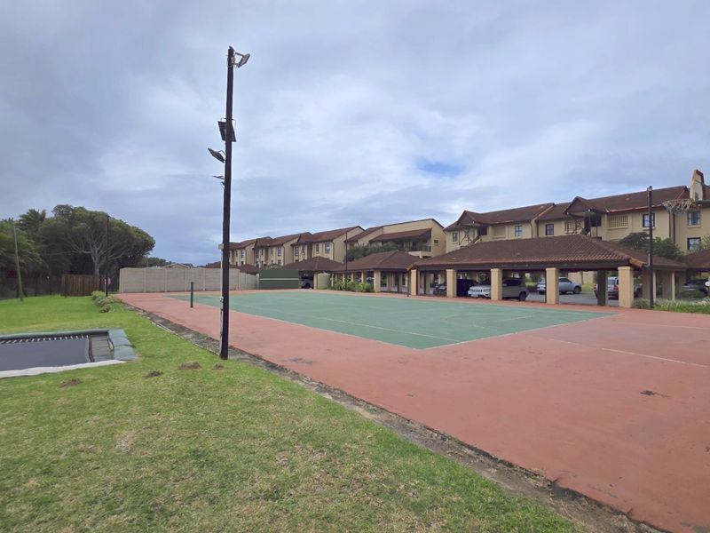 A communal sports facility featuring a multi-purpose court with basketball hoop and floodlighting, situated within an apartment complex with covered parking bays.