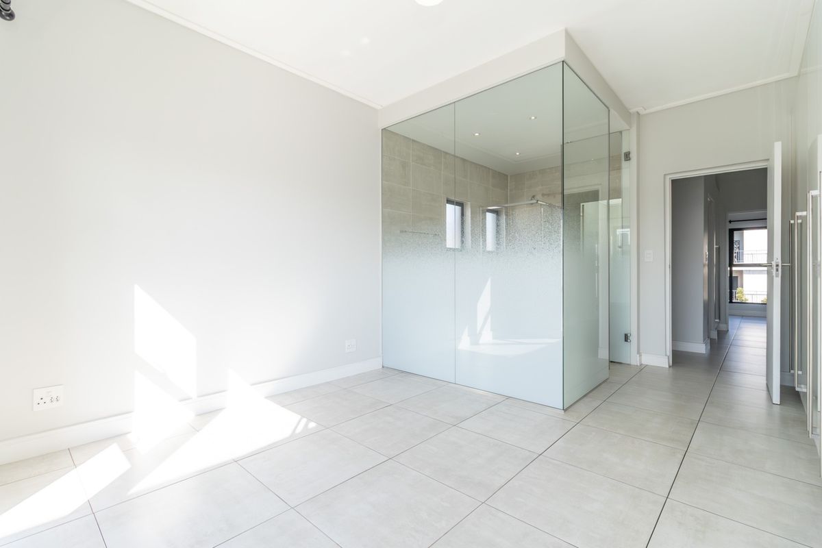A bright, modern bedroom featuring a sleek, glass-enclosed en-suite bathroom with tiled flooring and neutral finishes.