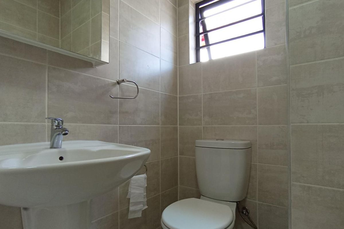 A compact, modern bathroom featuring neutral wall tiles, a pedestal basin, and a standard toilet with a small window for natural ventilation.