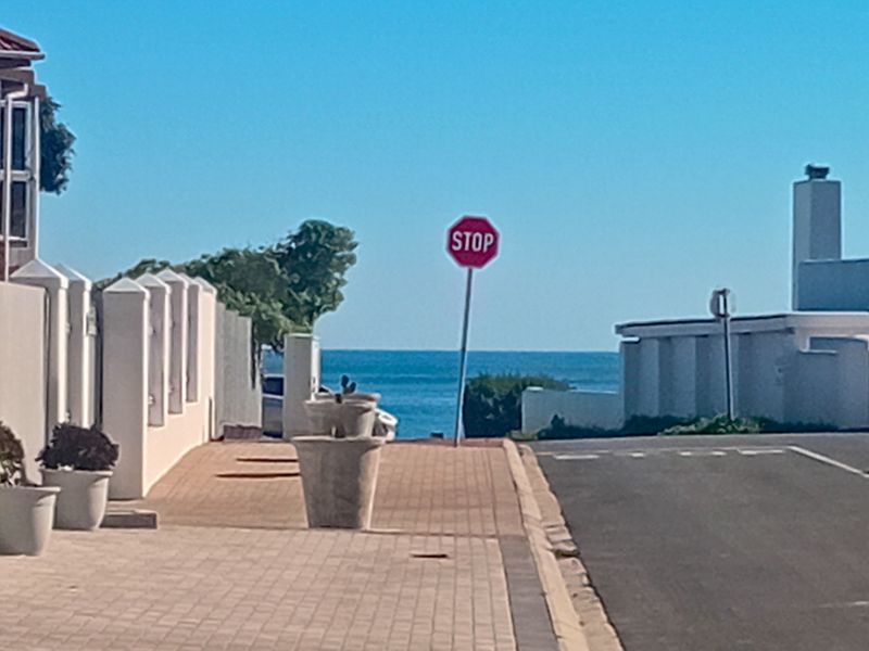 A street view showing a paved sidewalk and asphalt road leading towards a coastal horizon.