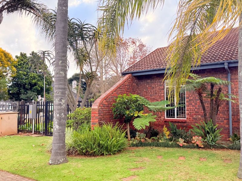 A view of the townhouse exterior featuring classic red brick facade, tiled roof, and established garden landscaping with mature palm trees.