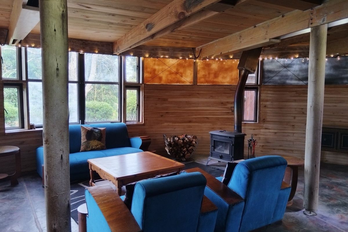 Rustic living room featuring exposed timber ceiling beams, wooden wall paneling, large windows, and a built-in wood-burning stove.