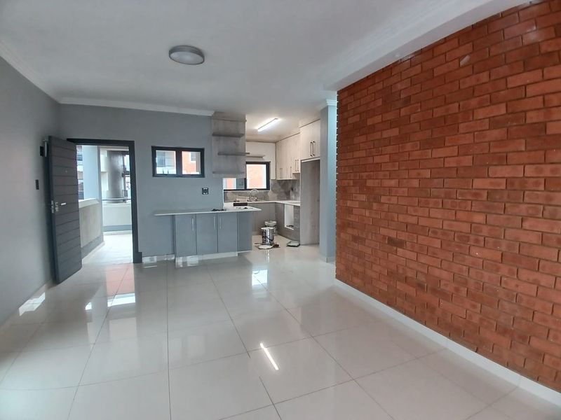 Spacious open-plan living and kitchen area featuring a striking exposed brick feature wall, modern cabinetry, and polished porcelain floor tiles throughout.
