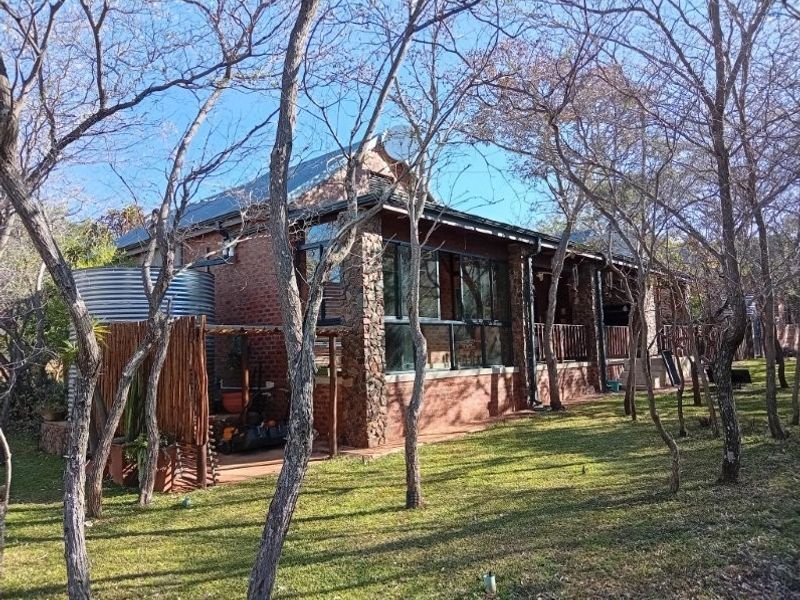 Rear view of a brick and stone retirement cottage featuring a covered patio, large windows, and an integrated water tank set within a wooded garden.