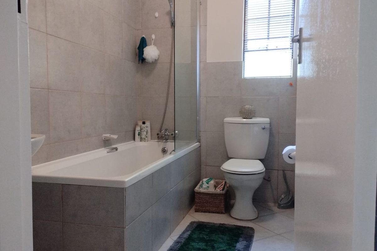 A functional bathroom featuring a tiled bathtub with a glass shower screen, a standard toilet, and a window with fitted blinds.